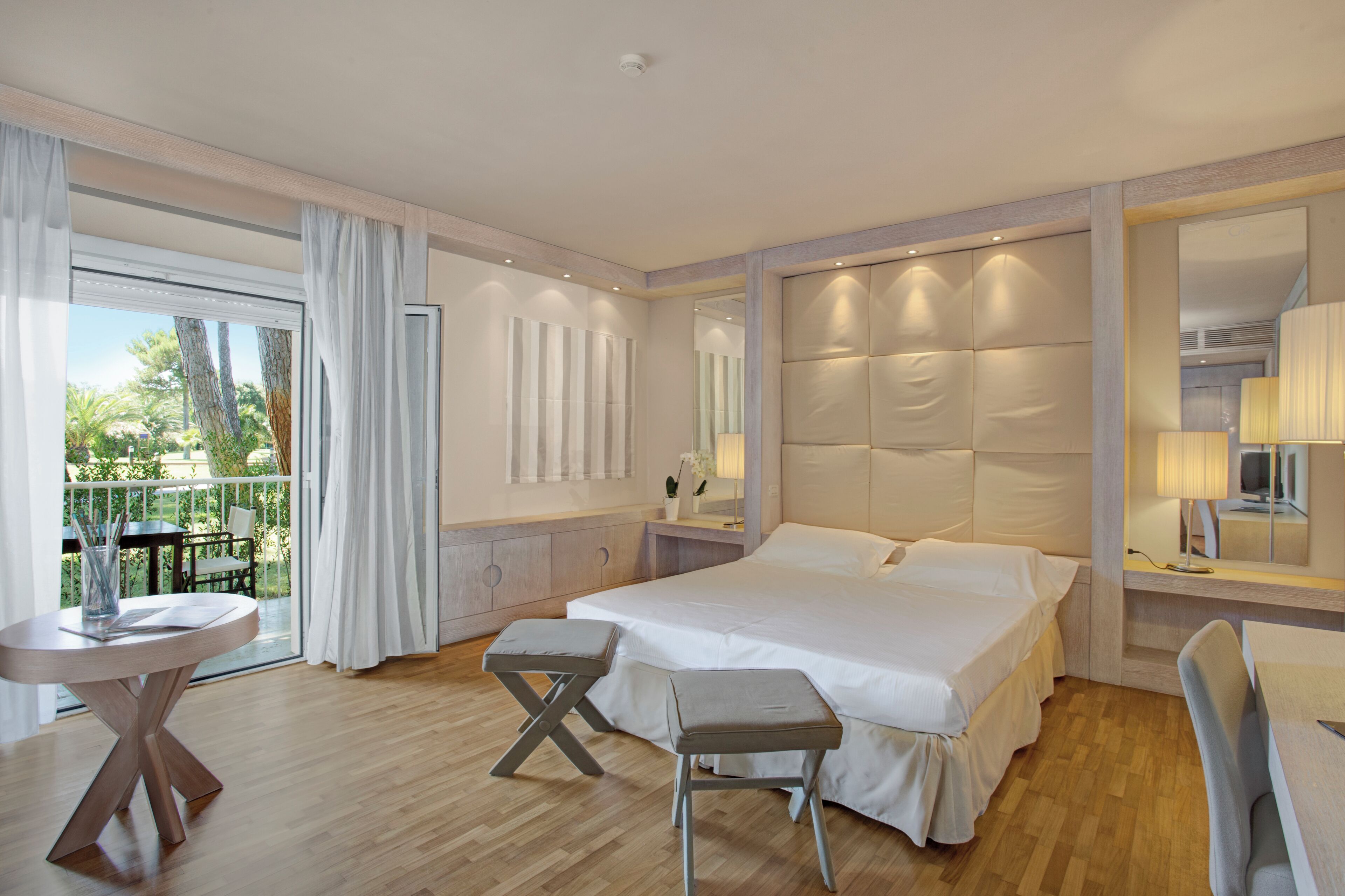 A hotel room with a large bed, a small table, a chair, a balcony with a table and chairs, and a large mirror.