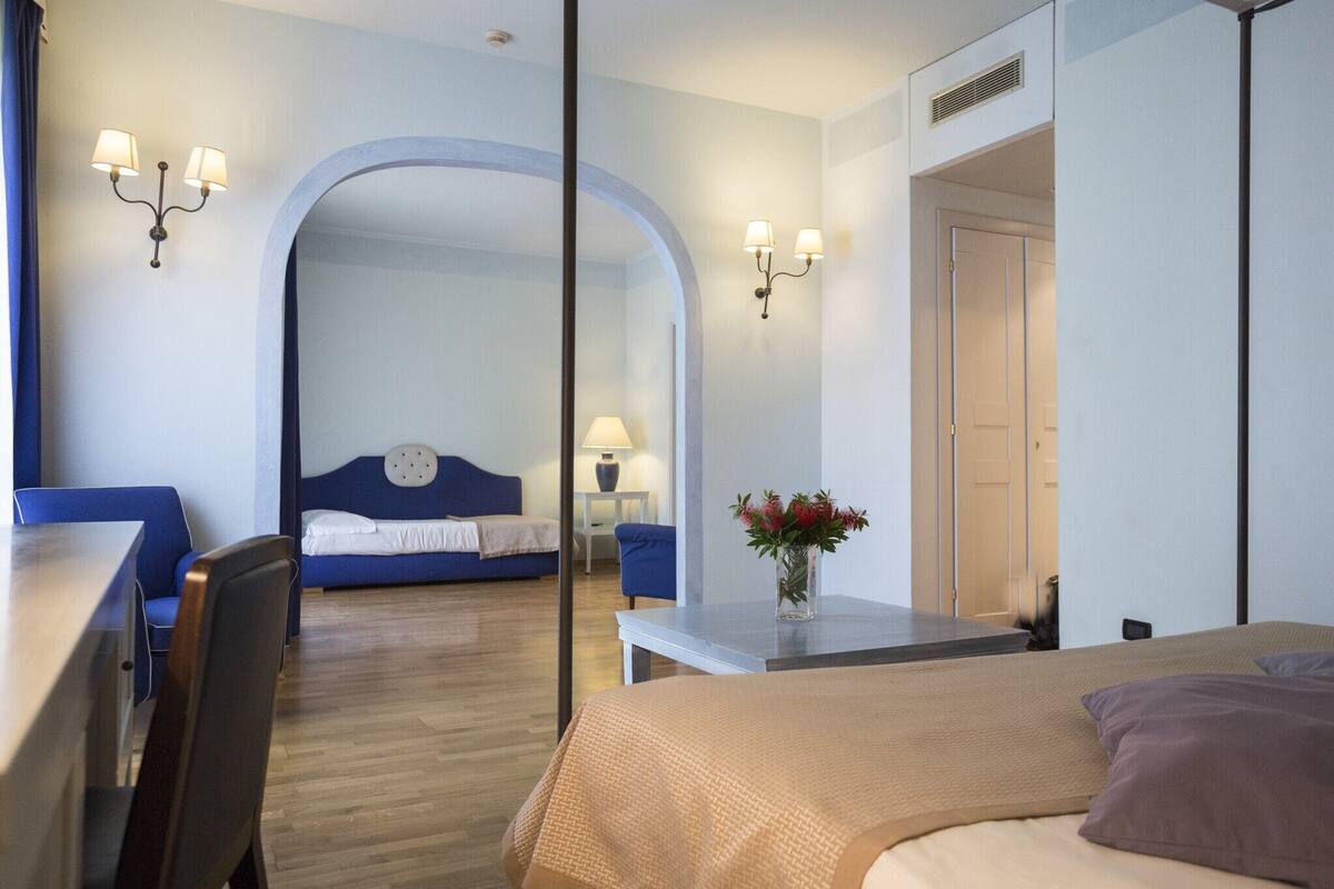 Une chambre d’hôtel avec un lit, un bureau, une chaise, une petite table ornée d’un vase de fleurs et un canapé bleu.