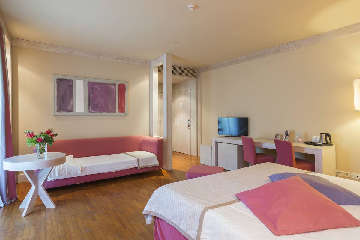 Une chambre d’hôtel avec un lit, un canapé rose, une petite table avec des fleurs, un bureau avec une chaise et un téléviseur.