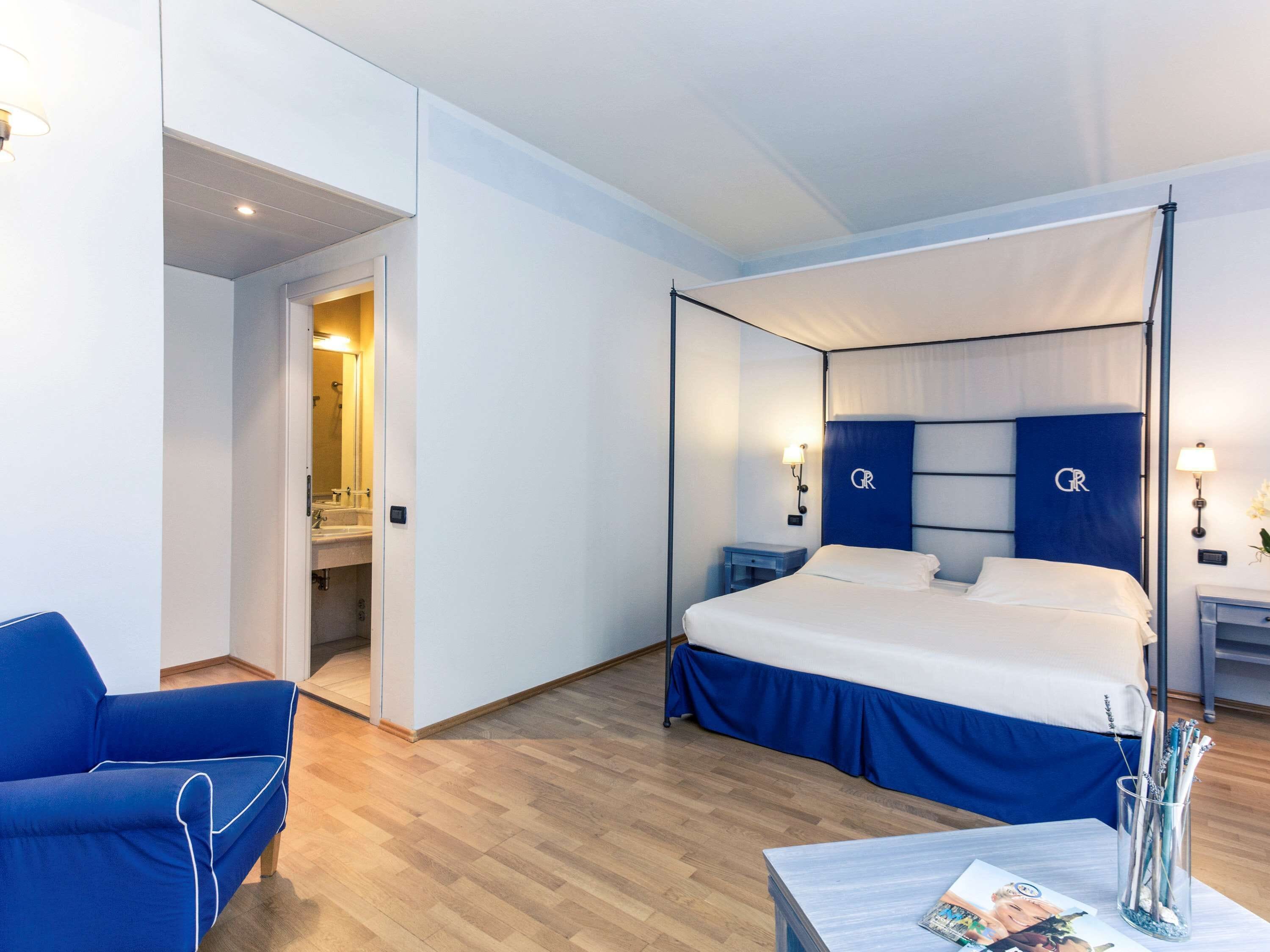 Een moderne hotelkamer met een groot bed, een zithoek met een blauwe bank, een klein tafeltje met een tijdschrift, en een badkamer zichtbaar door een geopende deur.