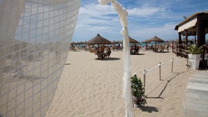 Ein Strand mit reetgedeckten Sonnenschirmen, einem sandigen Weg und einem eingezäunten Bereich mit Topfpflanzen.