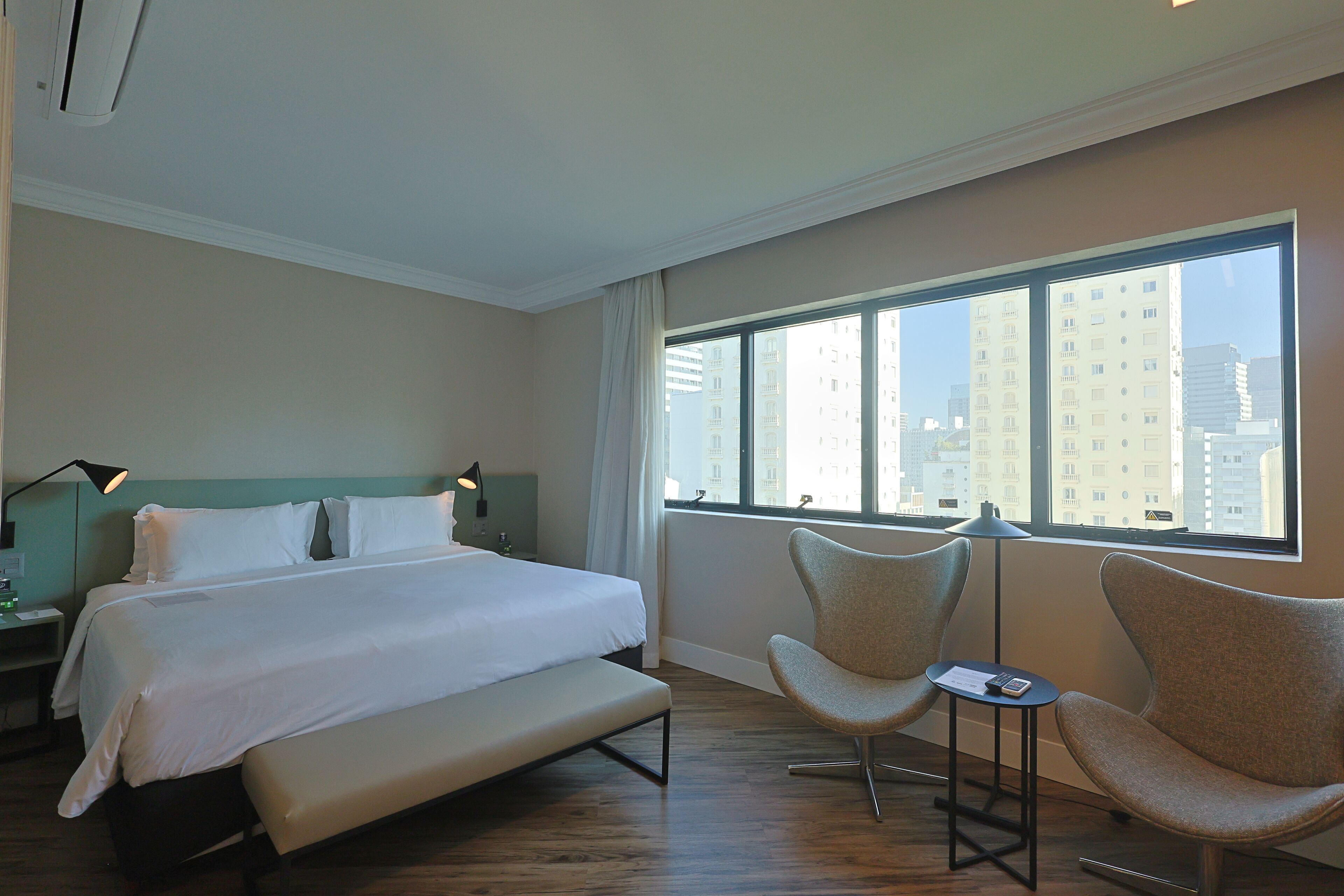 A hotel room with a large bed, two chairs, a small table, and a window with a city view.