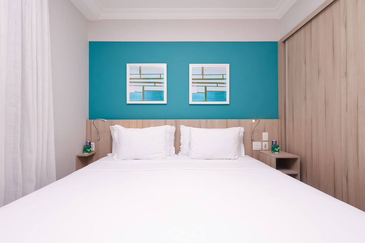 A hotel room with a large bed, two bedside tables, a nightstand, a teal accent wall, and two framed pictures.