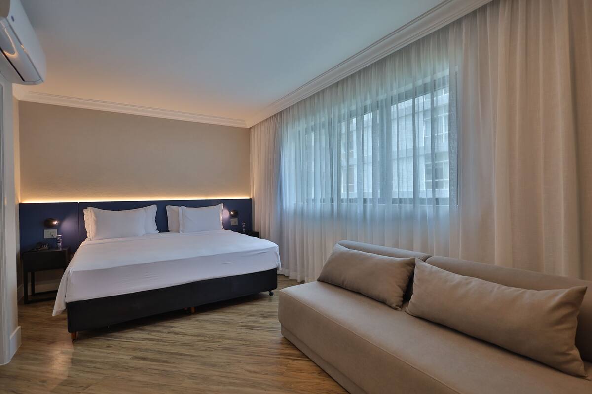 A hotel room with a bed, a sofa, a nightstand, and a window with sheer curtains.