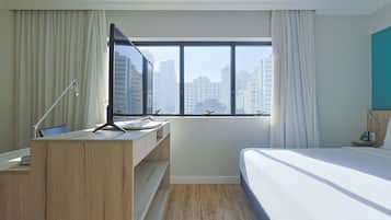Quarto de hotel com cama, uma secretária com computador, janela com cortinas e vista para edifícios da cidade.