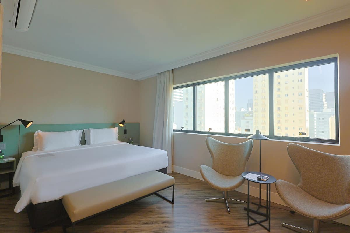 A hotel room with a large bed, two armchairs, a small table, and a window with a city view.