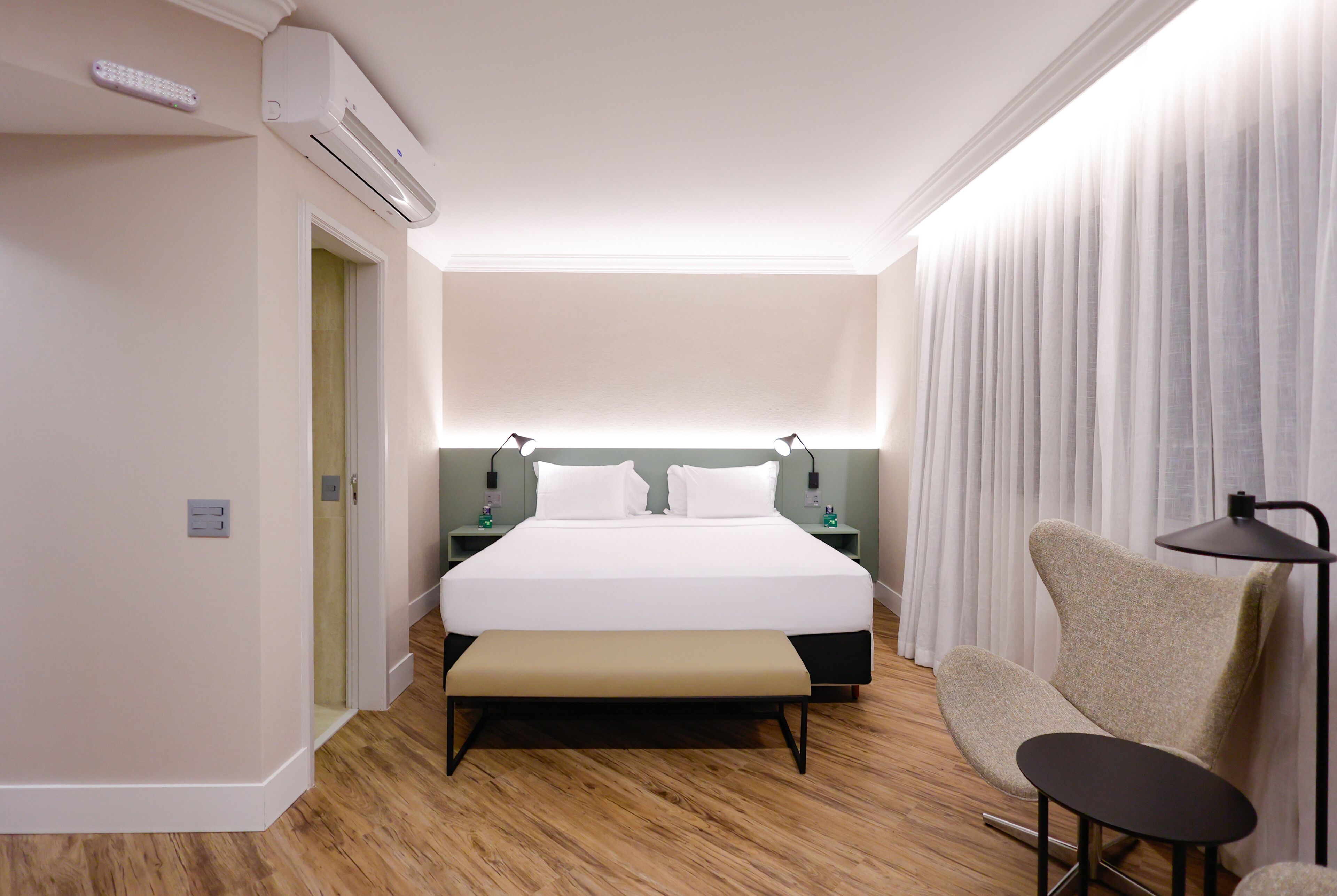 Quarto de hotel com cama, mesas de cabeceira, um banco, uma cadeira e uma luminária.