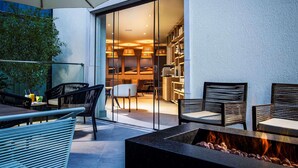 Un patio exterior con una fogata, una zona de estar y un ventanal que da a un espacio interior con estanterías y comedor.