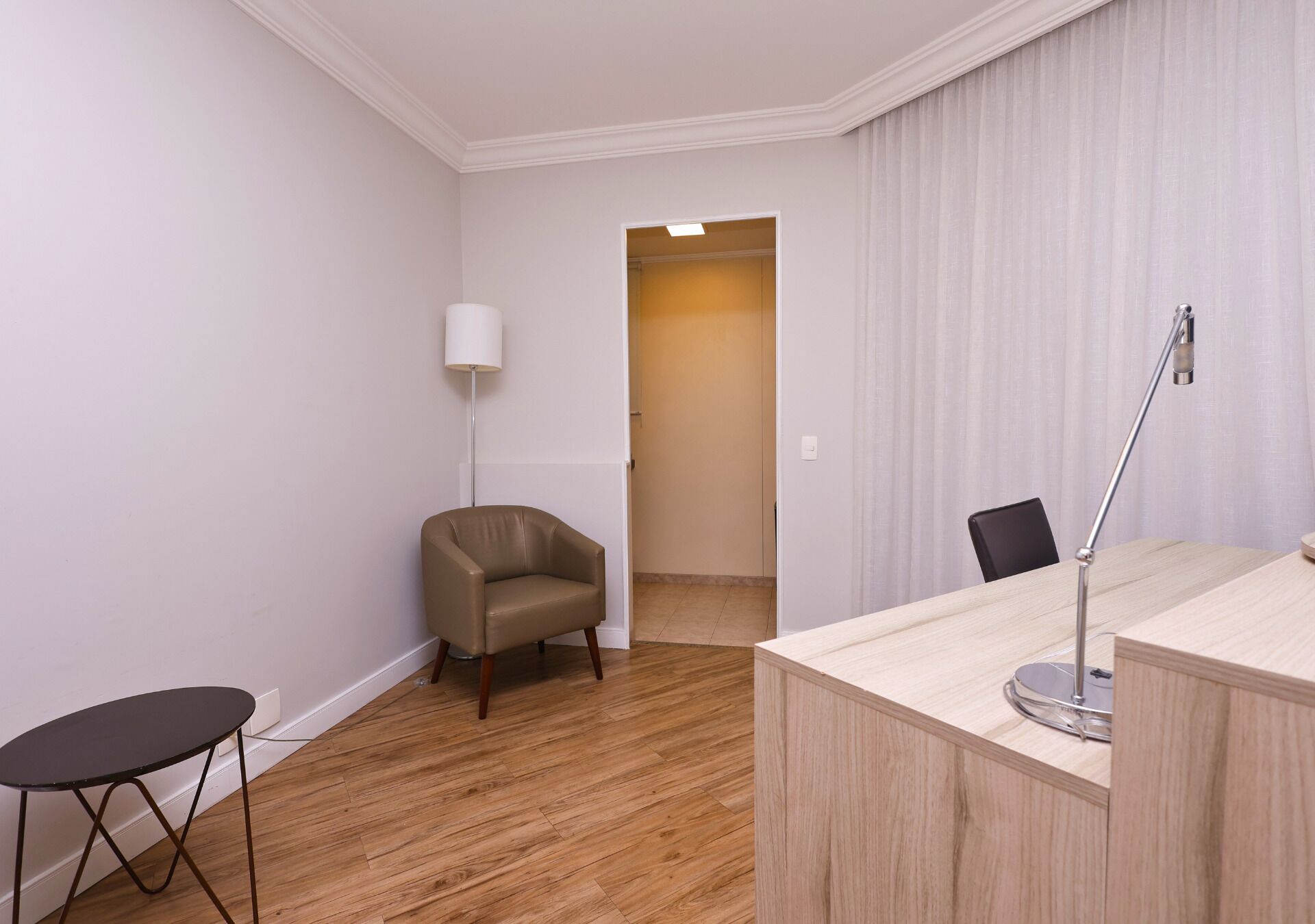 Quarto com piso de madeira, uma escrivaninha com luminária, uma cadeira e uma pequena mesa.