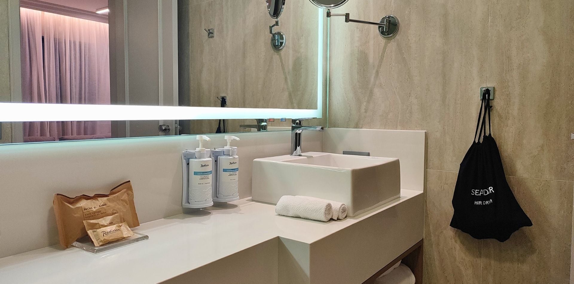 Casa de banho moderna com lavatório branco, espelho e toalha. Existem duas embalagens de loção e um saco de papel castanho no balcão.