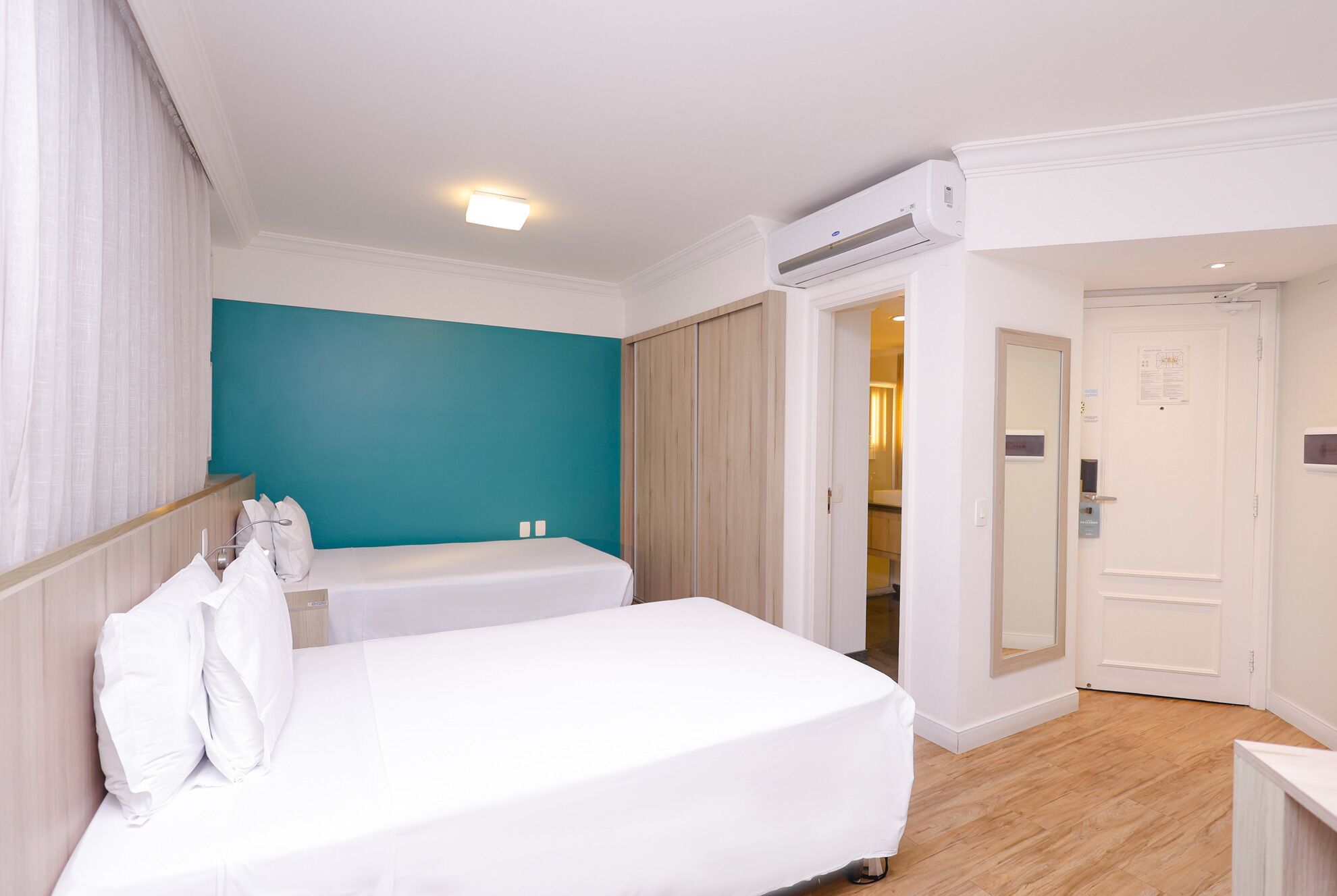 A hotel room with two beds, a teal accent wall, a wooden wardrobe, an air conditioning unit, and a door leading to a bathroom.