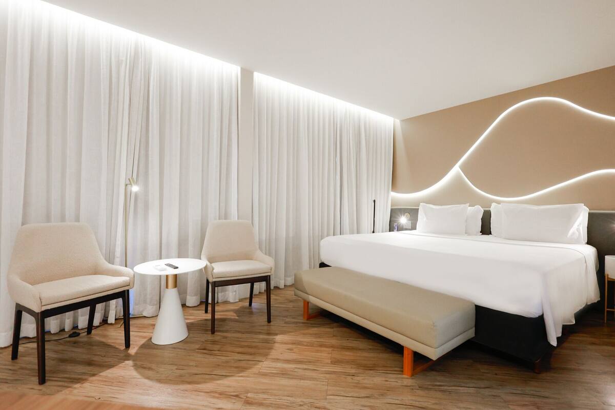 A modern hotel room with a large bed, two armchairs, a small round table, and a large window with sheer curtains.