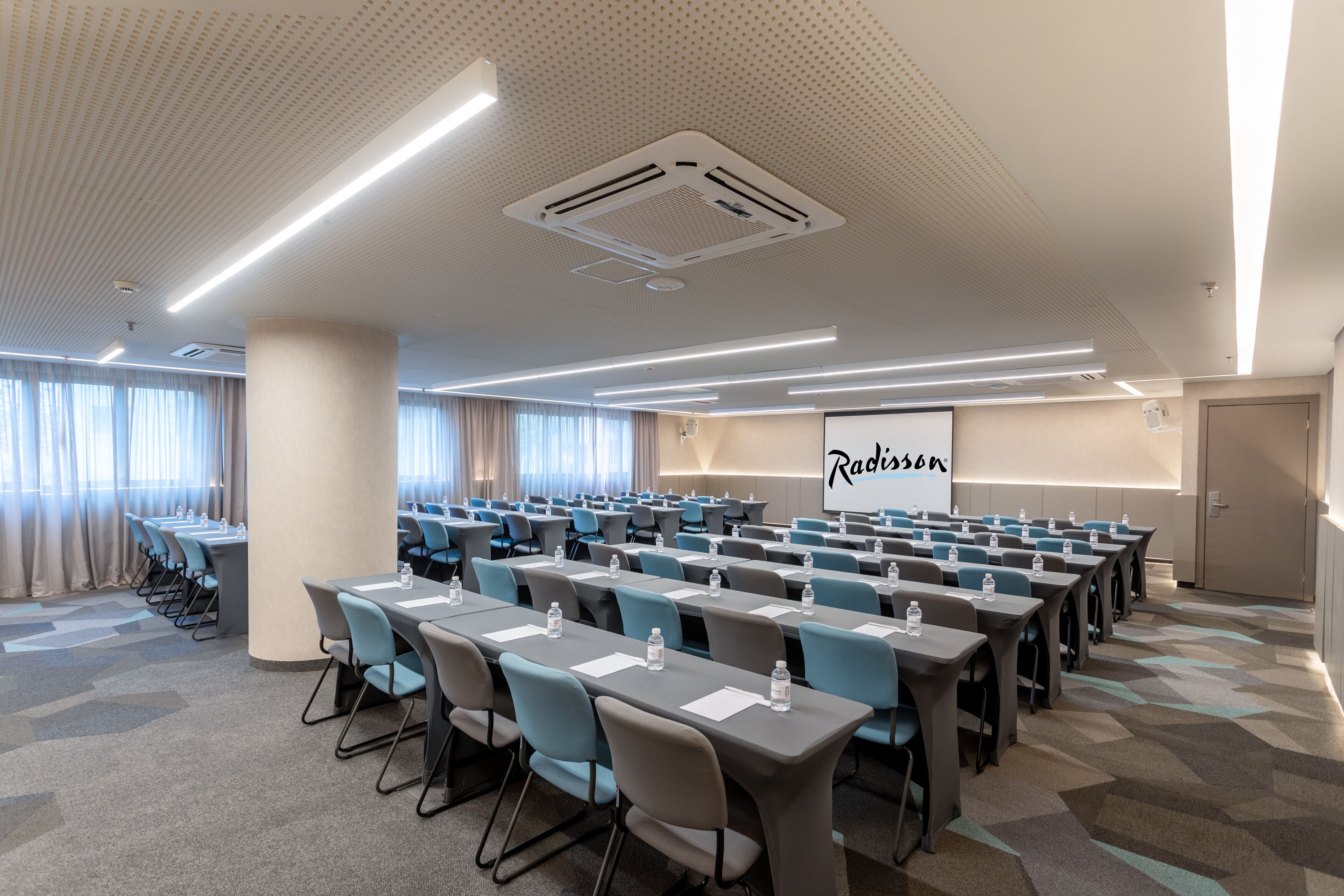 Un salón de conferencias con filas de mesas y sillas, una pantalla de proyector y un amplio ventanal con cortinas.