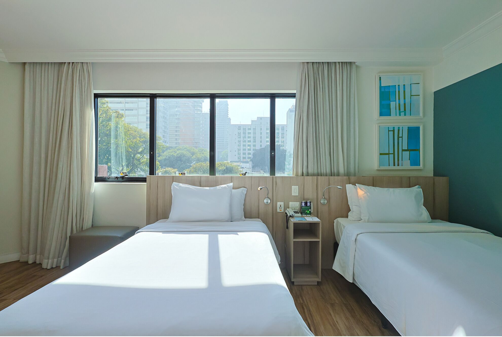 Habitación de hotel con dos camas, una mesita de noche, una ventana con cortinas y vistas a edificios.