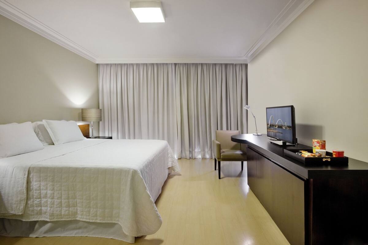 A hotel room with a bed, a desk with a television, a chair, and a window with curtains.