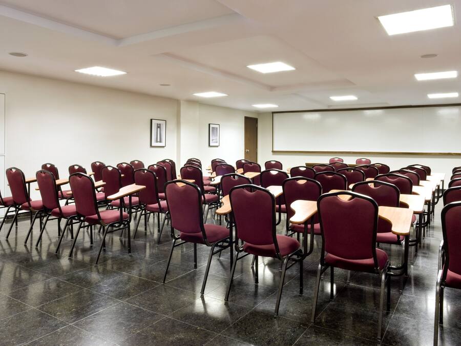 A conference room with rows of chairs and tables, a projector screen, and framed pictures on the walls.
