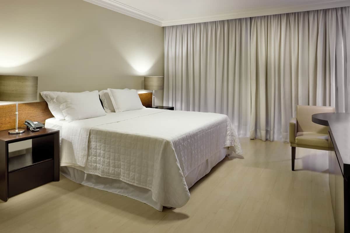 A hotel room with a large bed, a bedside table with a lamp, a chair, and a curtain.