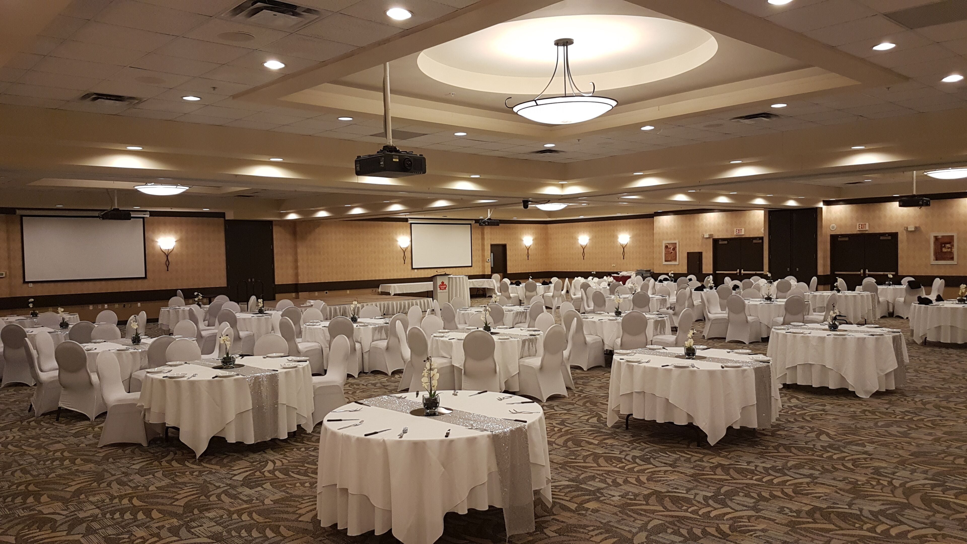 A large conference room with multiple round tables, white tablecloths, and chairs, equipped with a projector and screen.
