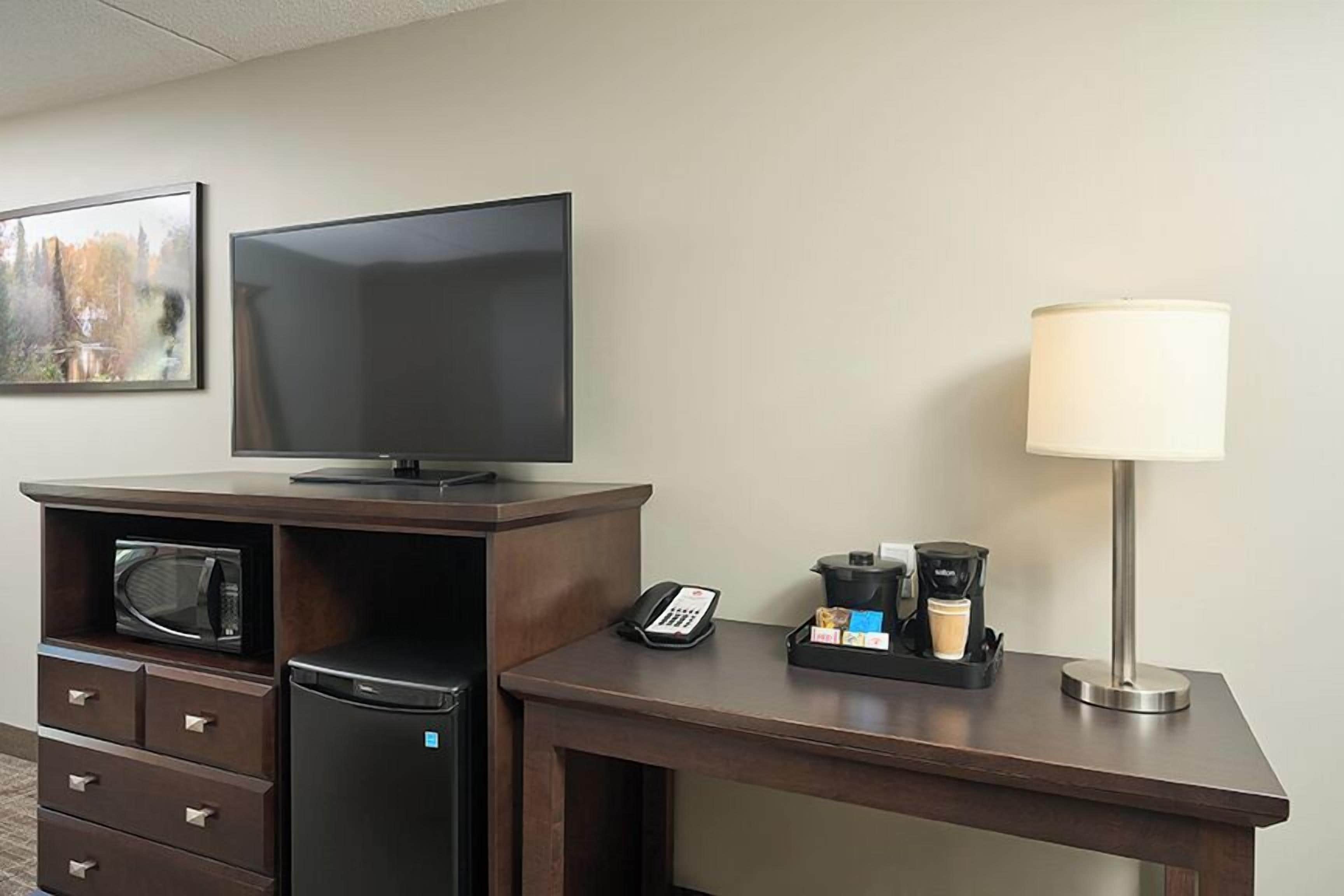 A hotel room with a flat-screen TV, a microwave, a refrigerator, a desk with a lamp, a phone, and a coffee maker.