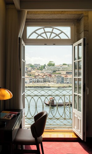 A room with a view of a river, a chair, a desk with a lamp, and a window with a metal railing.