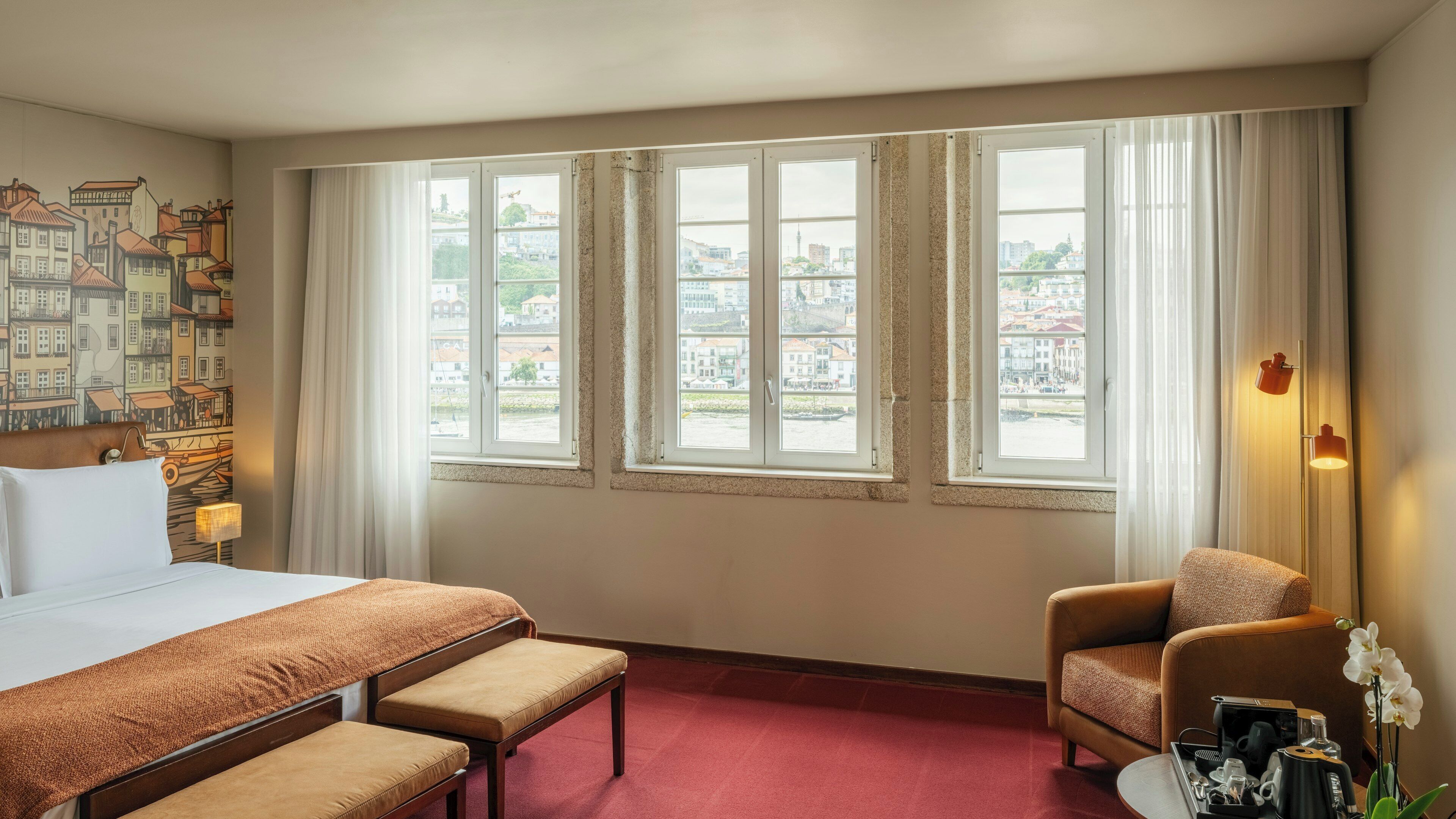 Une chambre d’hôtel avec un lit, deux chaises, une petite table avec une machine à café, et une vue sur les bâtiments à travers de grandes fenêtres.