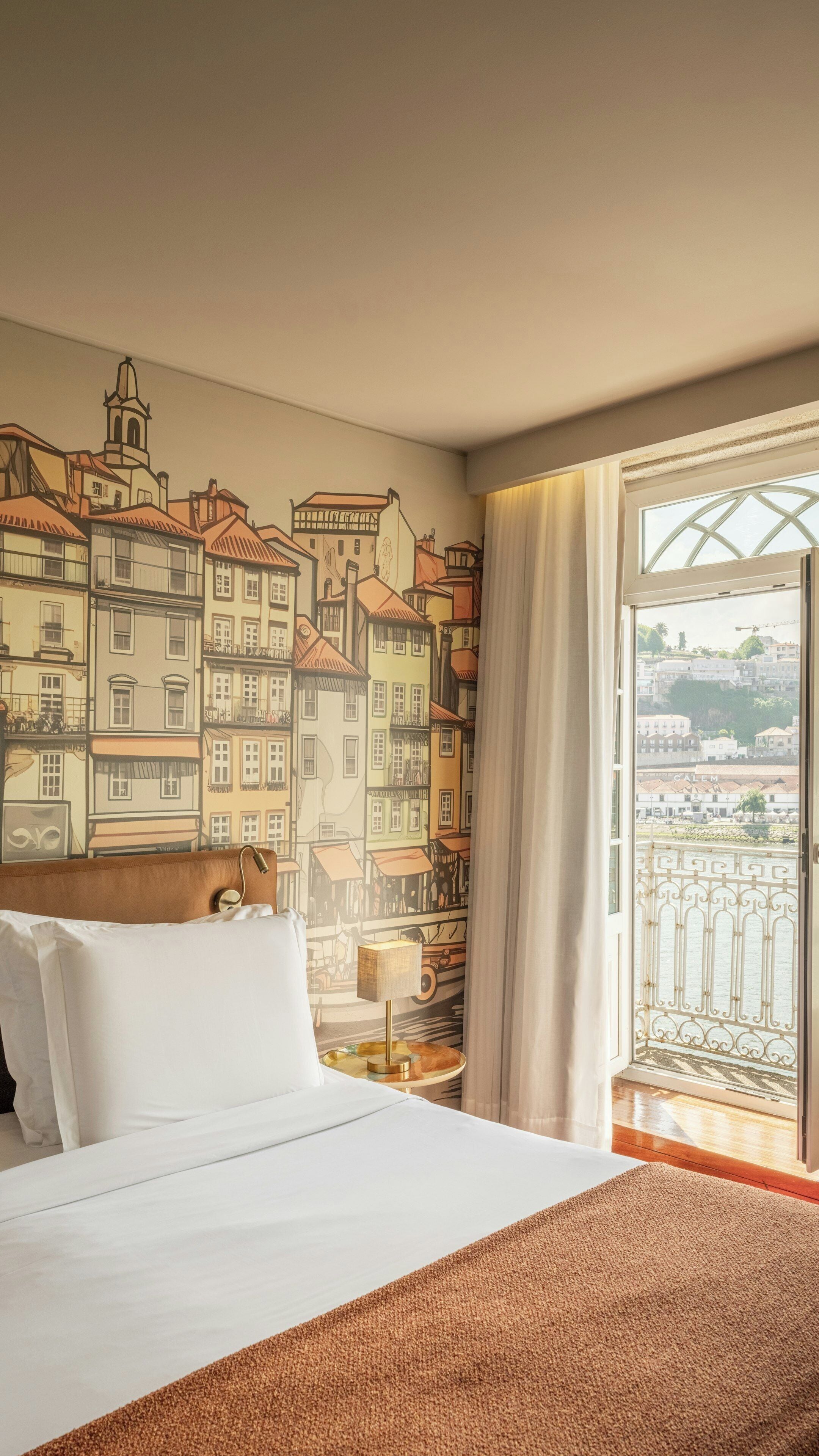 Una habitación de hotel con cama, mesita de noche, lámpara, ventana con cortinas y un mural de edificios.