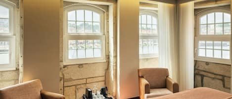 A room with a bed, two armchairs, a small table with a tray, and large windows with views of a cityscape.