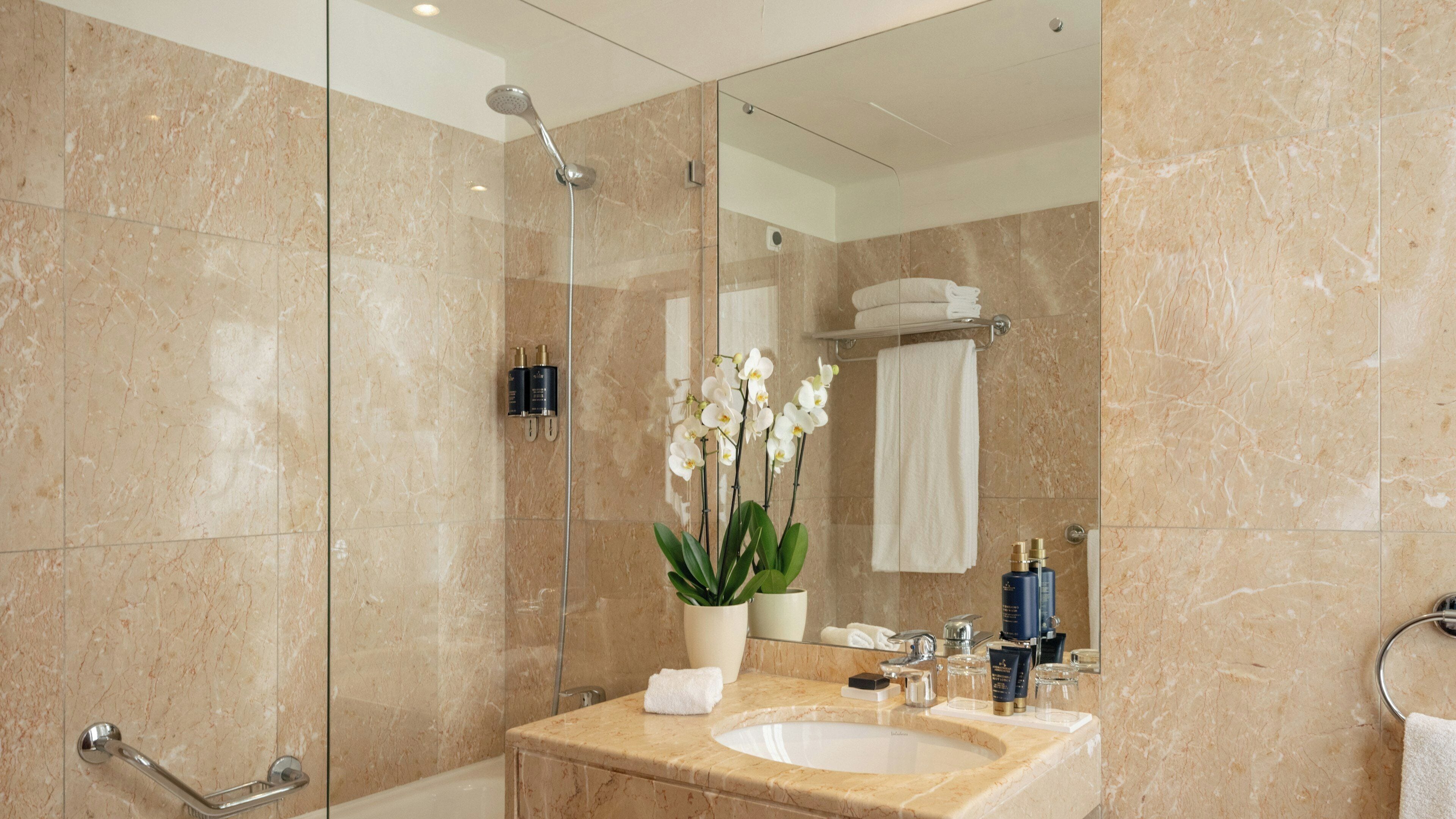 A modern bathroom with a glass shower enclosure, marble walls, a sink with toiletries, and a potted orchid.