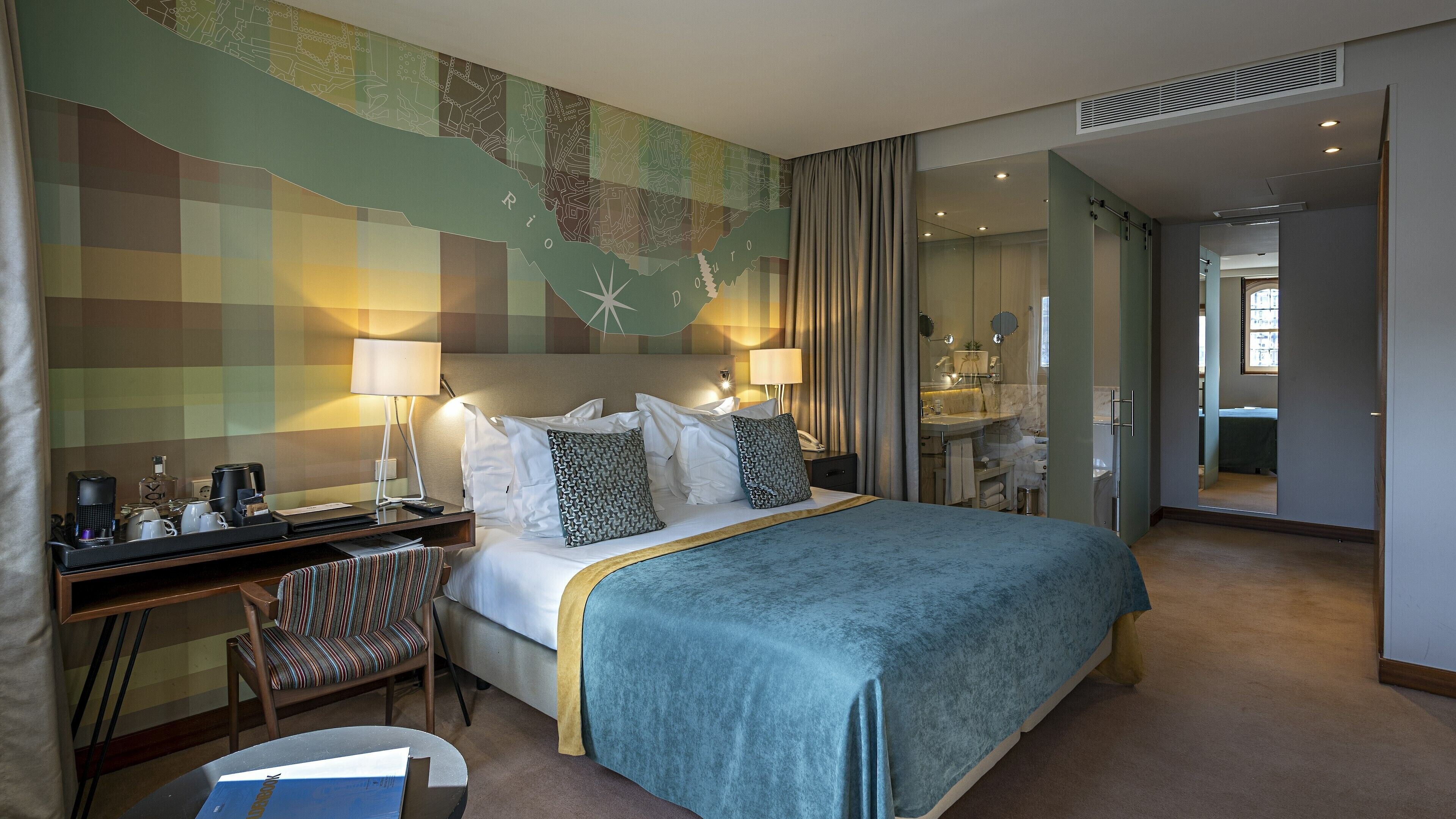 Ein Hotelzimmer mit einem Bett, Nachttischlampen, einem Schreibtisch mit Stuhl, einem Spiegel und einem durch eine offene Tür einsehbaren Badezimmer.