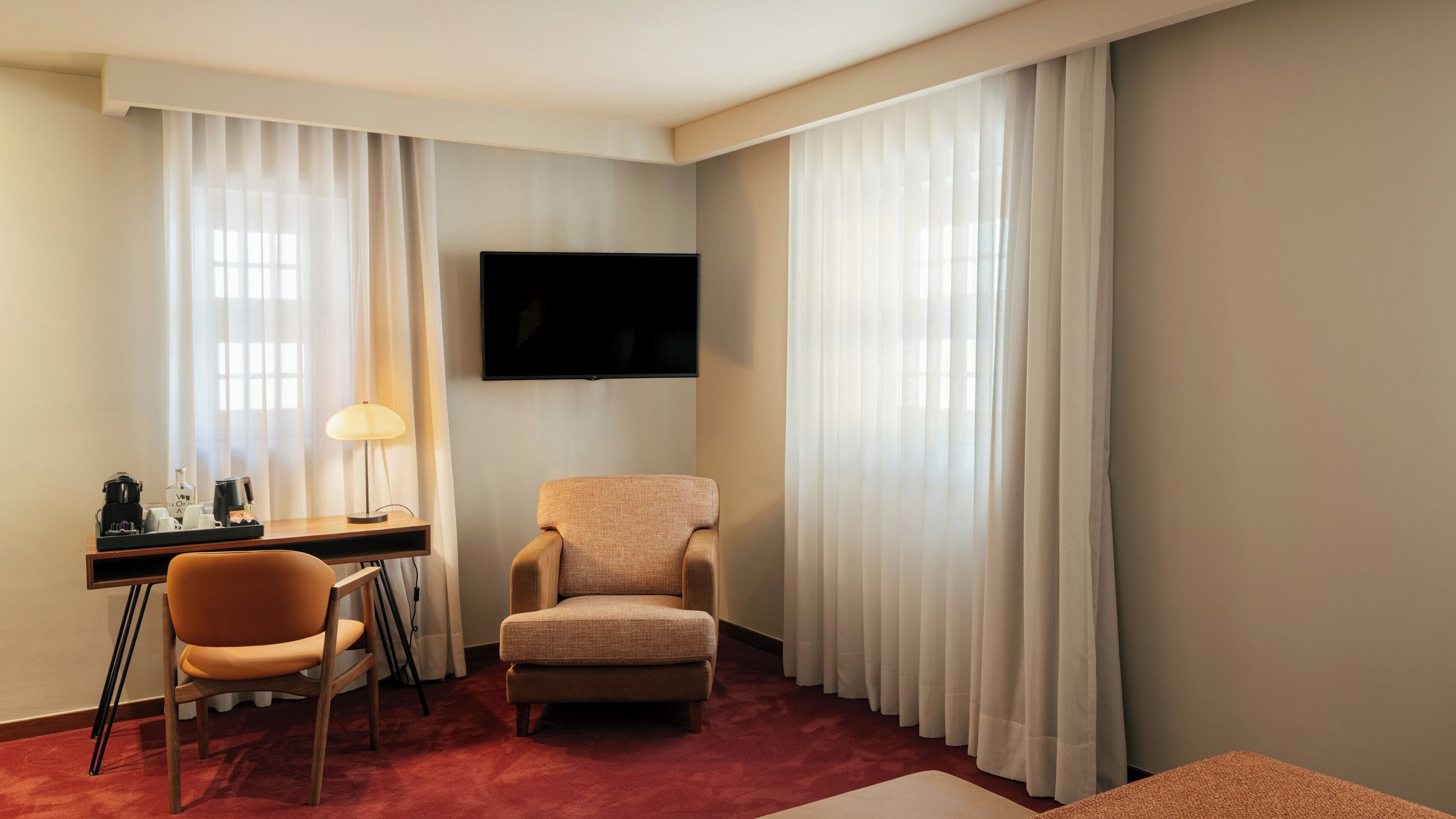A hotel room with a desk, chair, armchair, TV, and large windows with curtains.