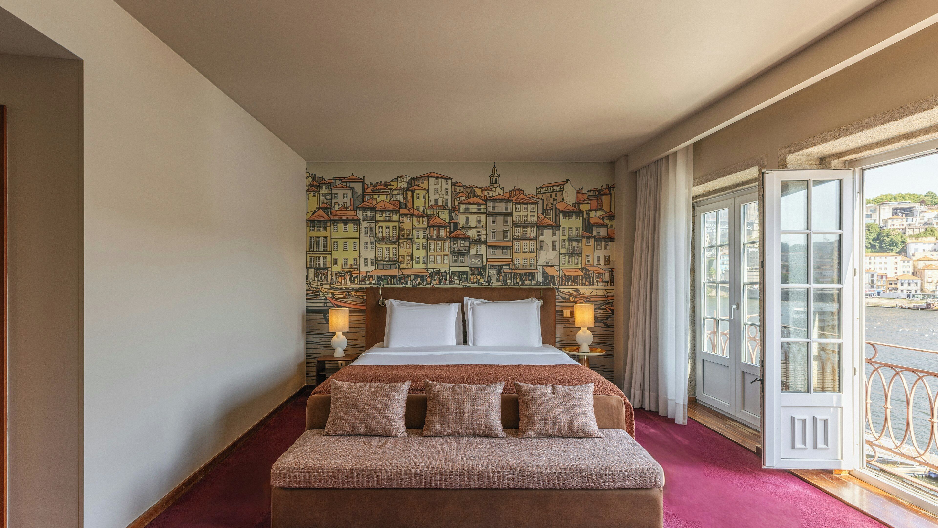 A hotel room with a bed, a bench, a cityscape mural, a window with a view, and a door.