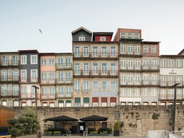 A row of multi-story buildings with balconies, a stone wall in the foreground, and outdoor seating with umbrellas.
