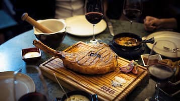 Un canard rôti, présenté sur une planche en bois avec une fourchette à découper, entouré de verres à vin et d’assiettes sur une table.