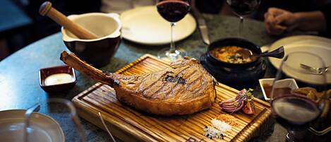 A roasted duck on a wooden board with a carving fork, surrounded by wine glasses and dishes on a table.