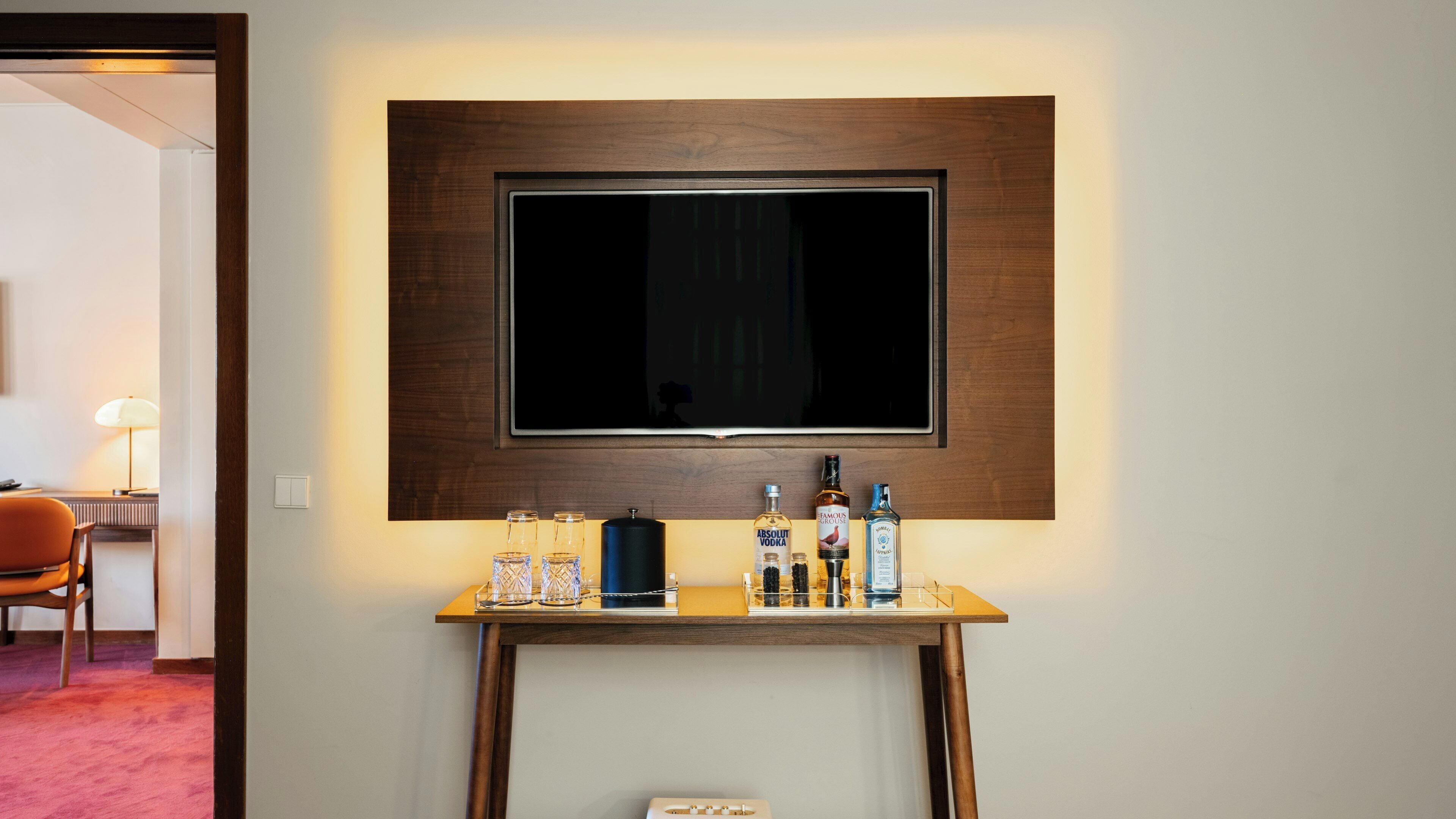 A hotel room with a mounted flat-screen TV, a small wooden console table with various bottles, and a chair.