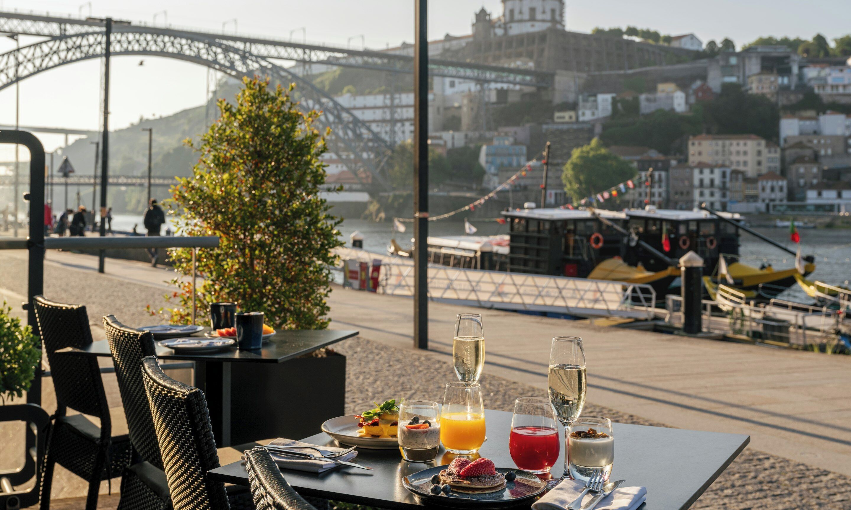 A table with breakfast items and drinks, overlooking a river with boats and a bridge.