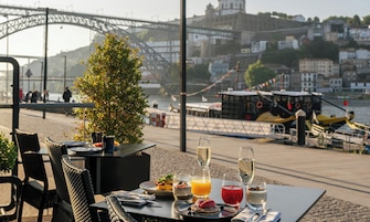 Mesa com itens de café da manhã e bebidas, com vista para um rio com barcos e uma ponte.