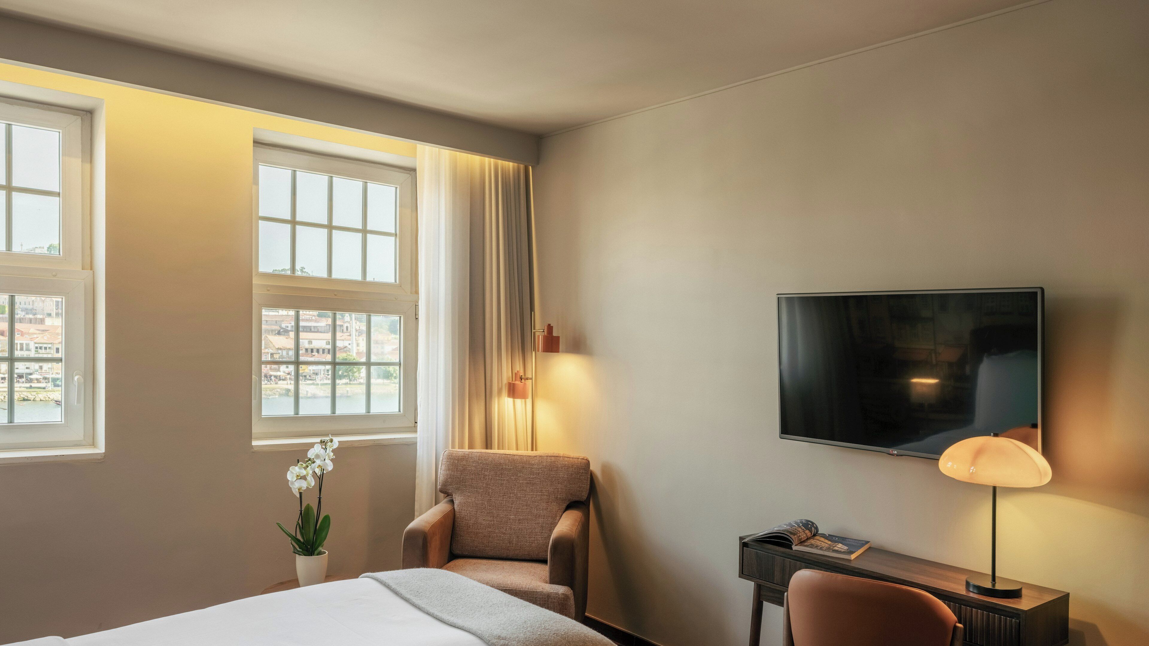 Ein Hotelzimmer mit Bett, Stuhl, Schreibtisch mit Lampe, Fernseher und Fenster mit Vorhängen.
