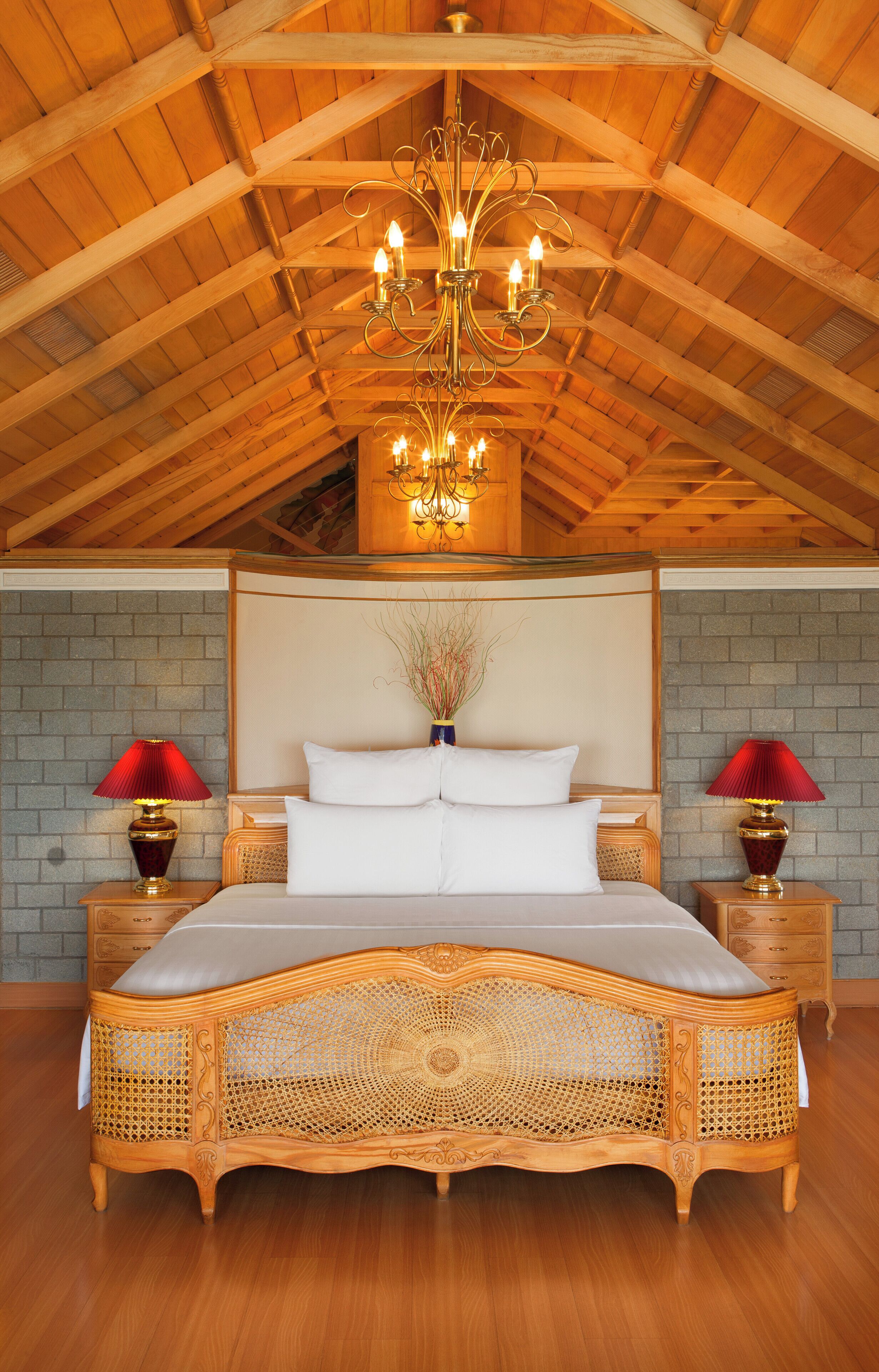 A large bed with a wooden frame and a woven headboard, two bedside tables with lamps, a chandelier, and a high wooden ceiling.