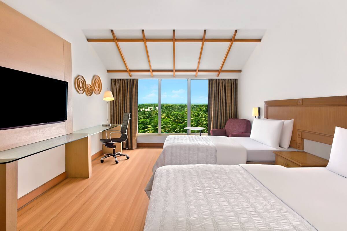A hotel room with two beds, a desk with a TV, a small table, and a view of greenery through large windows.
