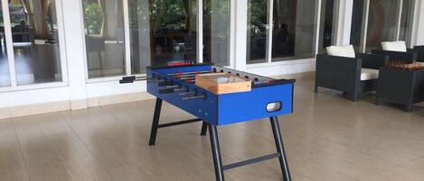 A foosball table in a room with large windows, a ceiling fan, and a seating area with a table and chairs.