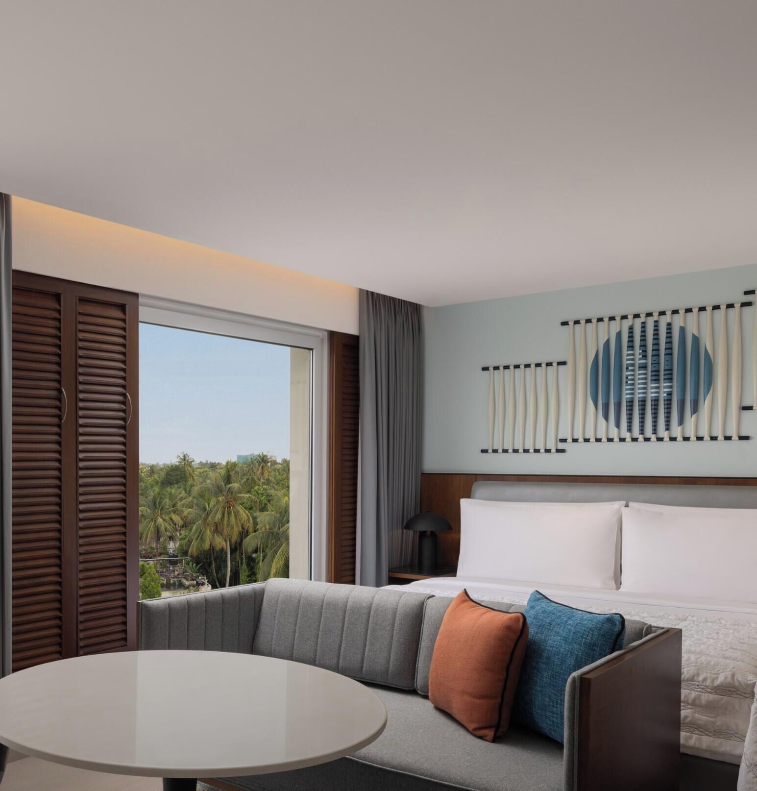Quarto de hotel com sofá, mesa redonda, grande janela com persianas e vista para palmeiras, além de uma obra de arte na parede.