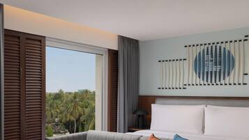 Quarto de hotel com sofá, mesa redonda, grande janela com persianas e vista para palmeiras, além de uma obra de arte na parede.