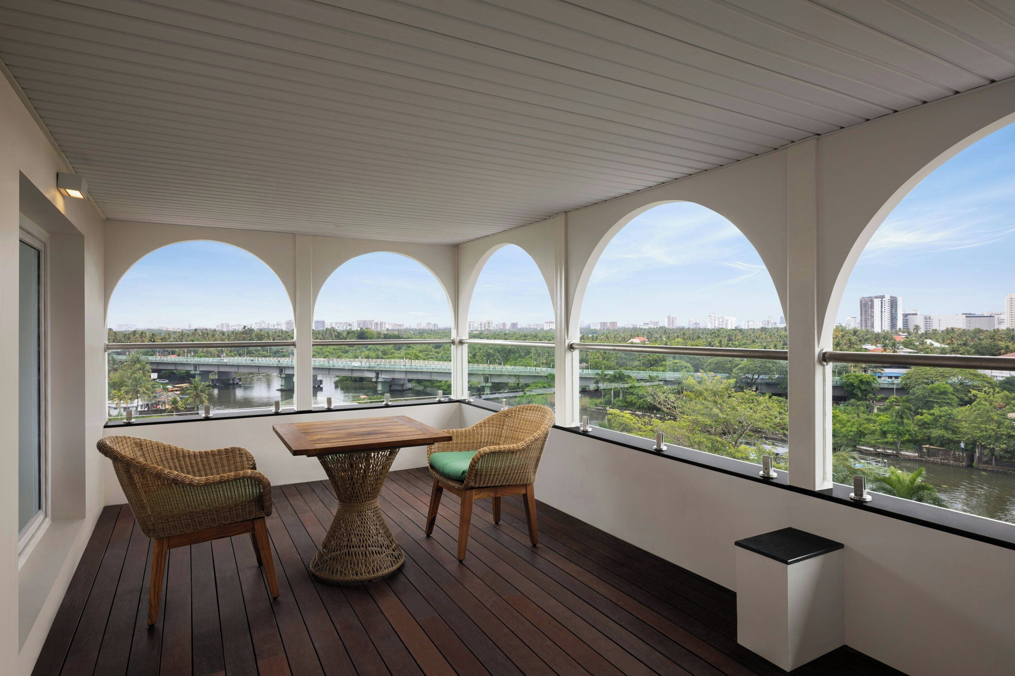 Un balcón con una mesa de madera y sillas de mimbre, con vistas a un paisaje urbano y un río.