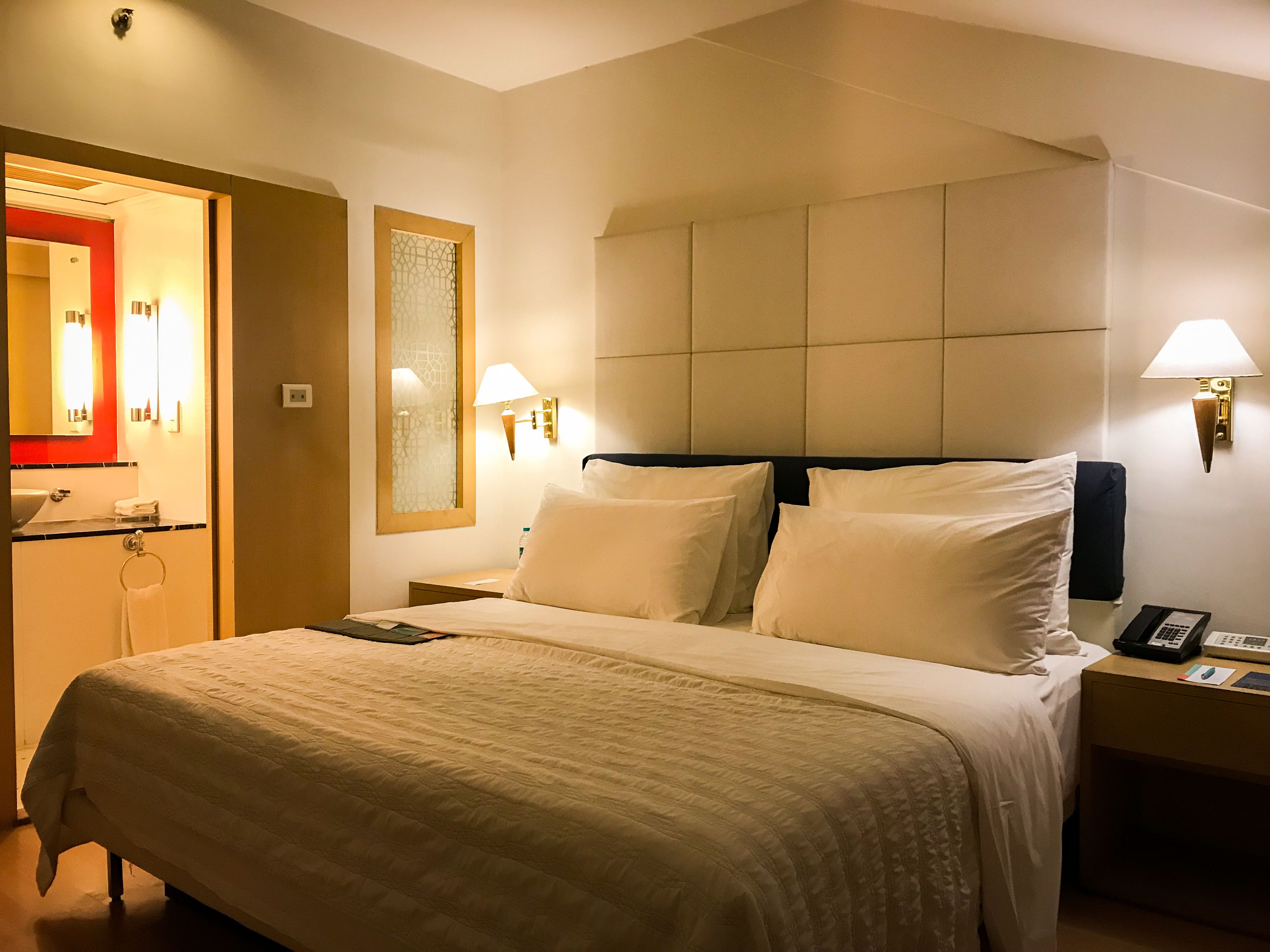 A hotel room with a large bed, two bedside tables, a nightstand, a mirror, and a wall-mounted light fixture.