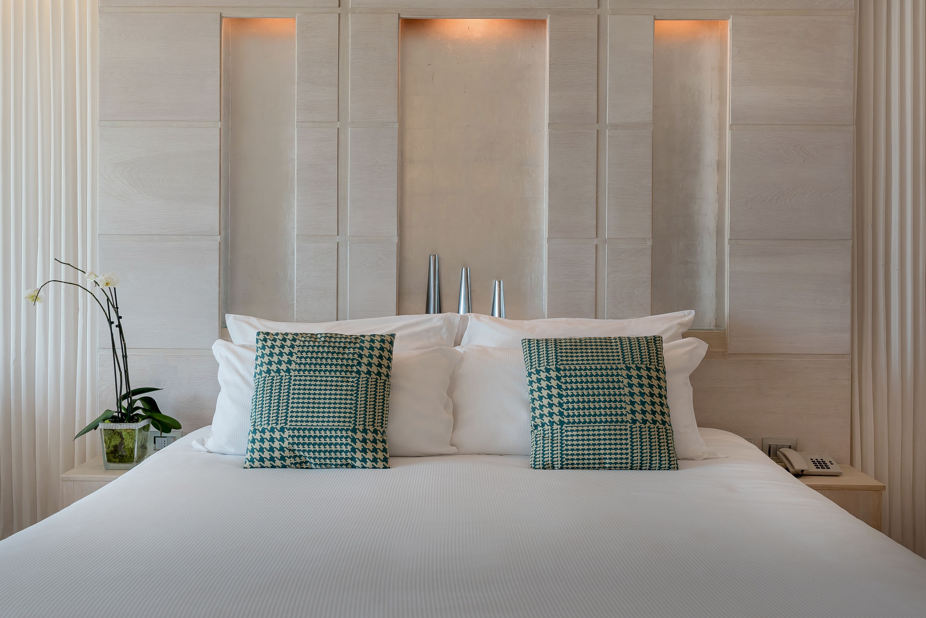 Una cama bien hecha con sábanas blancas y dos almohadas estampadas, una planta en la mesita de noche y un teléfono.