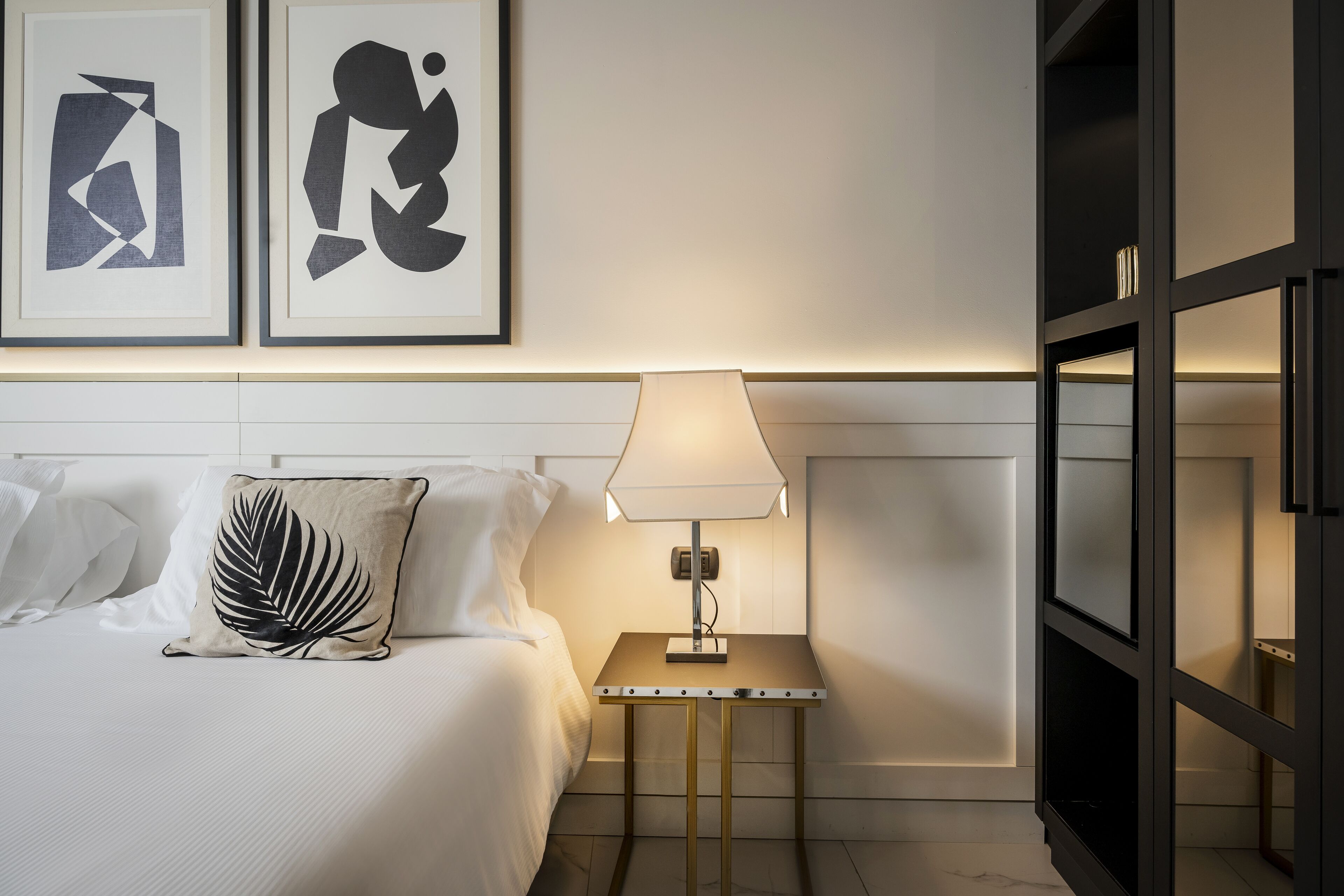 Una cama bien tendida con sábanas blancas y un cojín decorativo, una mesita de noche con lámpara, y dos obras de arte abstracto en la pared.