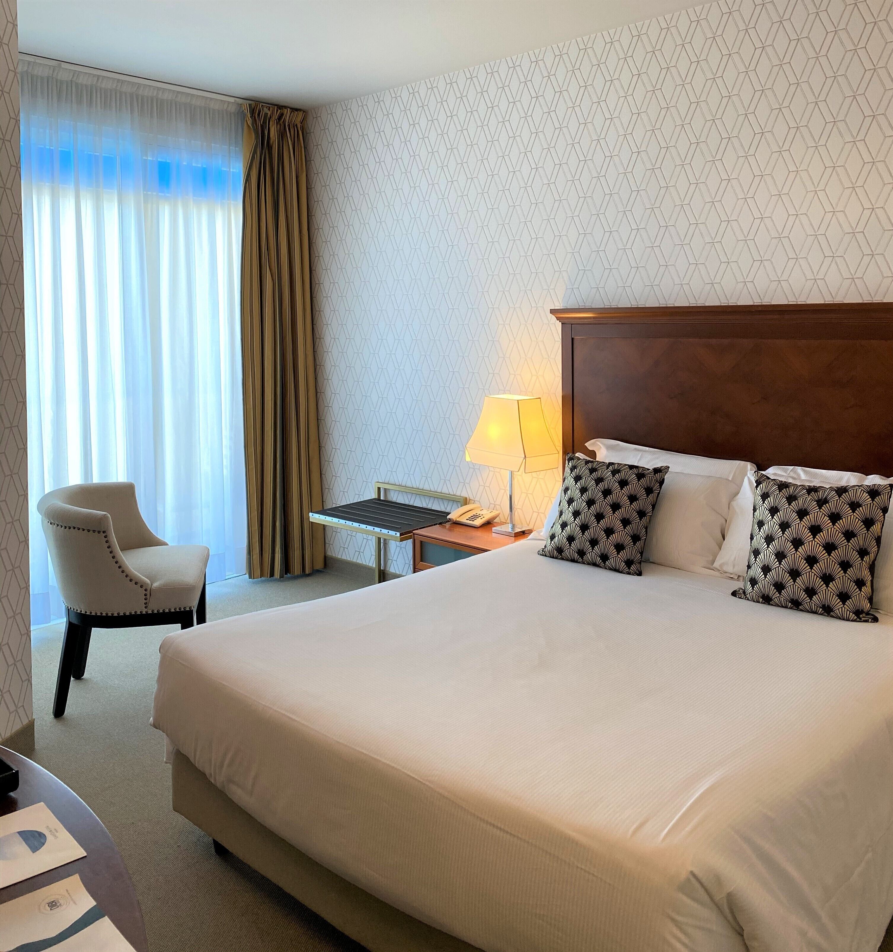 A hotel room with a large bed, a chair, a desk with a lamp, and a patterned wall.