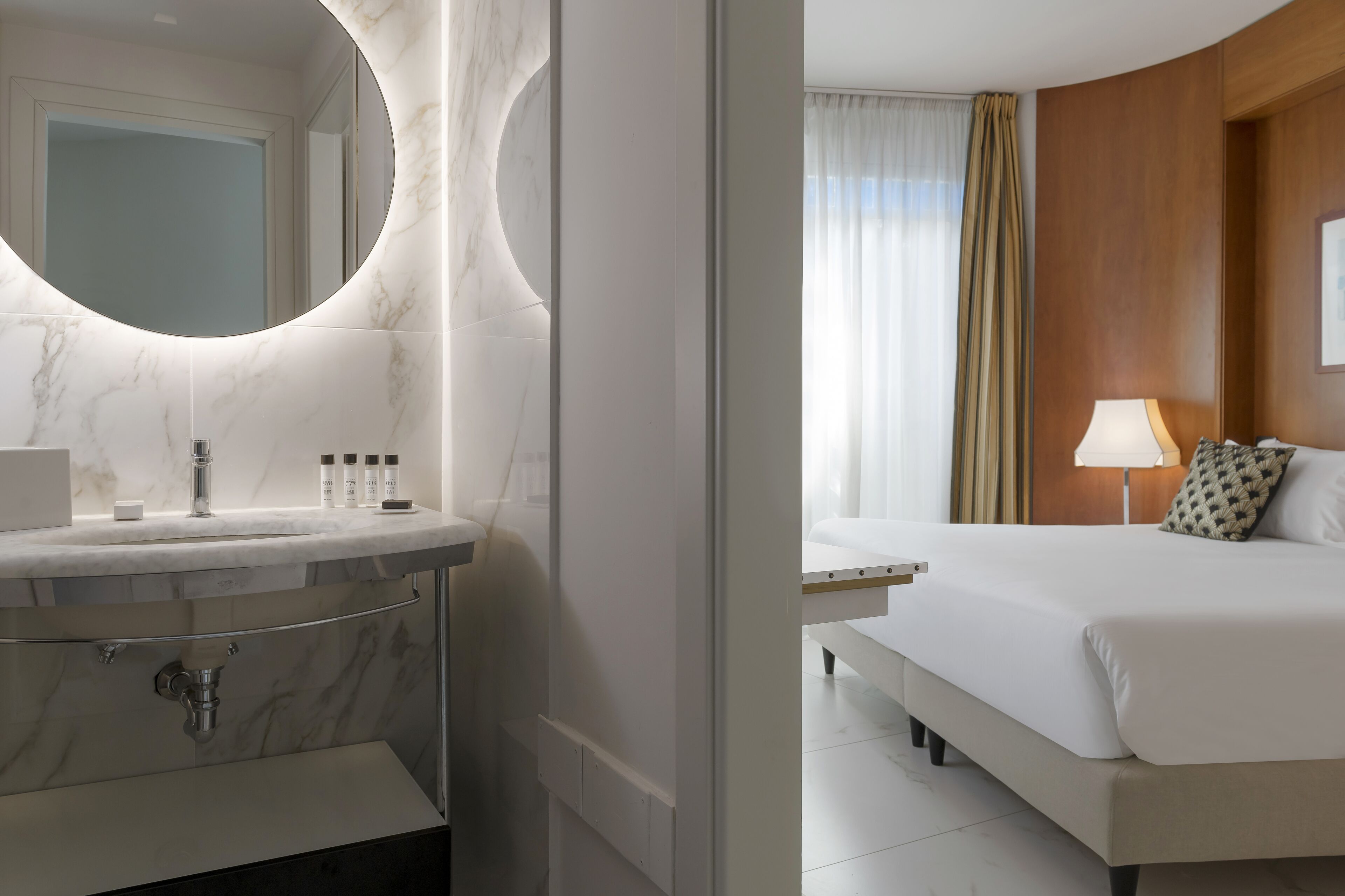 A modern bathroom with a marble vanity, a large mirror, and a bed with a wooden headboard in the adjacent room.