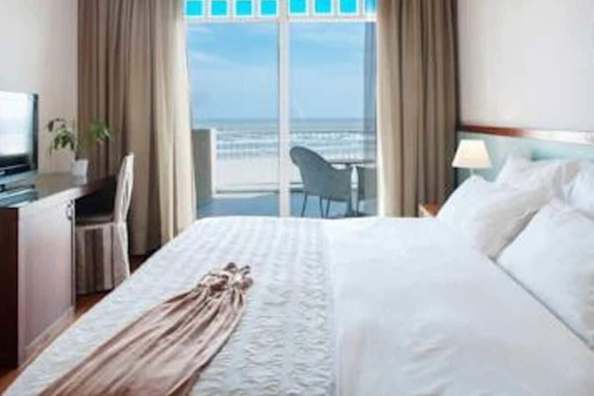 A hotel room with a bed, a desk, a chair, a lamp, and a view of the ocean through a large window.